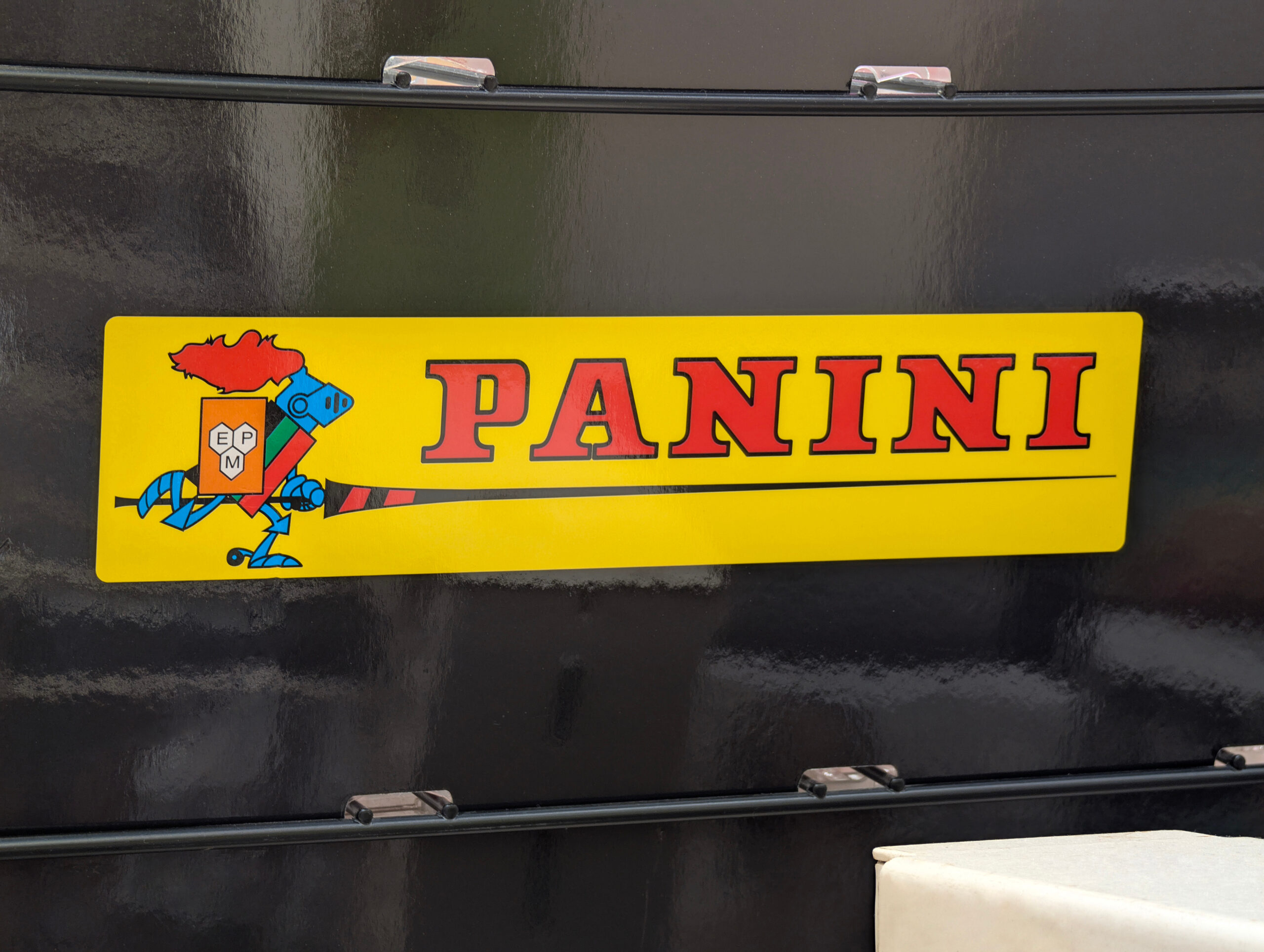Bright yellow license plate reading PANINI with a cartoon crest on the left, mounted on a dark vehicle.