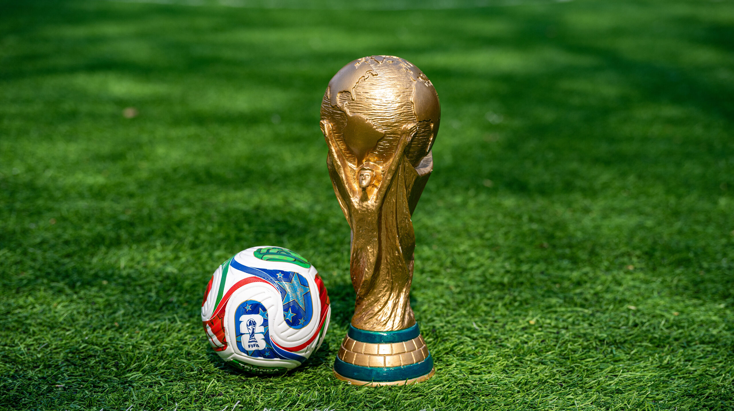 Golden World Cup trophy standing upright on a green soccer field beside a colorful soccer ball