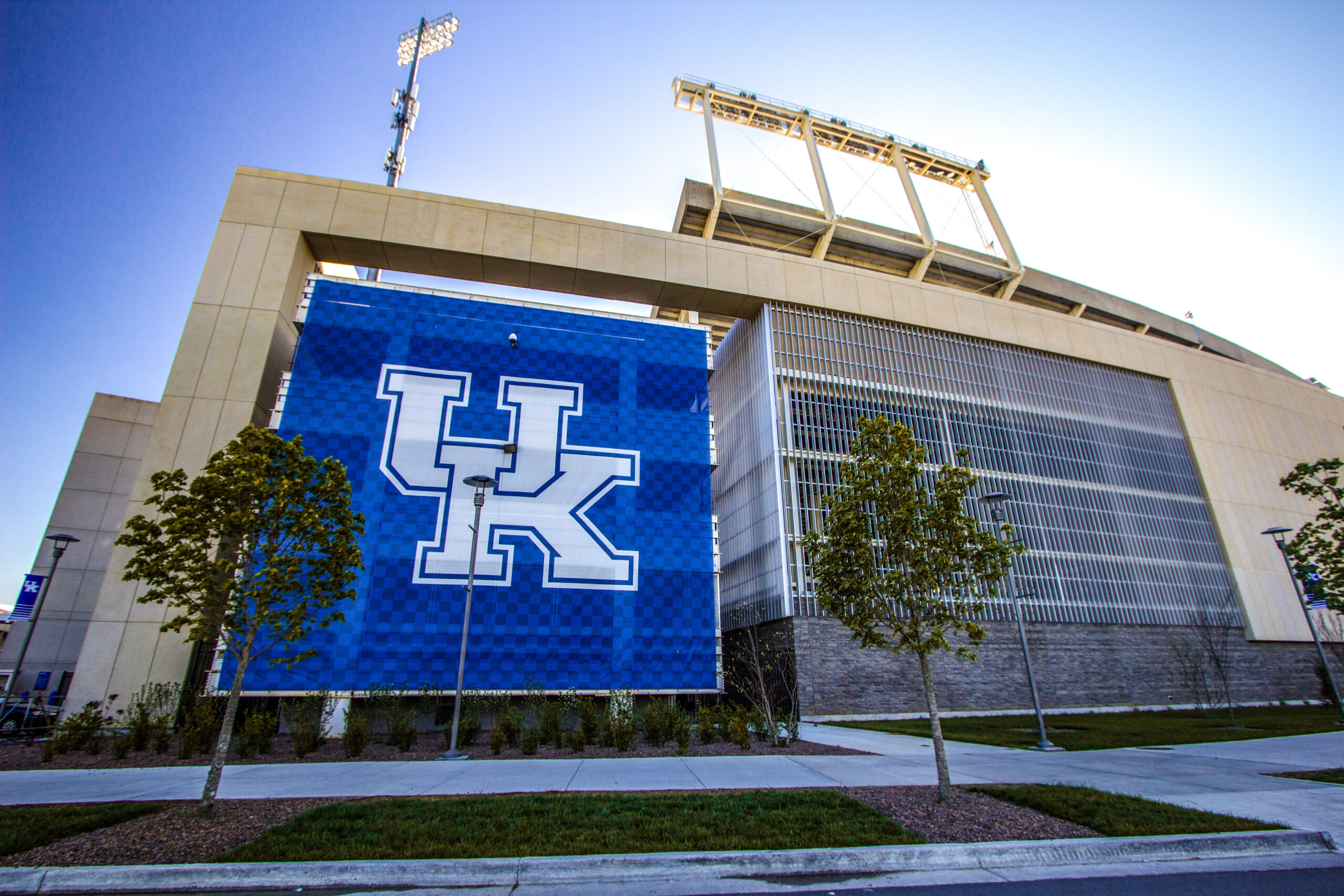 Exterior view of a modern stadium with a large blue UK banner displaying the university's white 'UK' logo, flanked by trees.