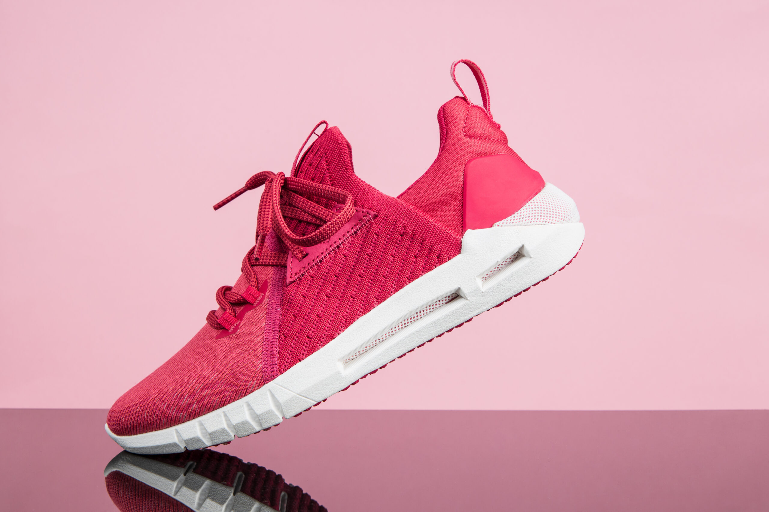 Pink athletic sneaker with a white sole angled against a pink background, showing laces and knit upper.