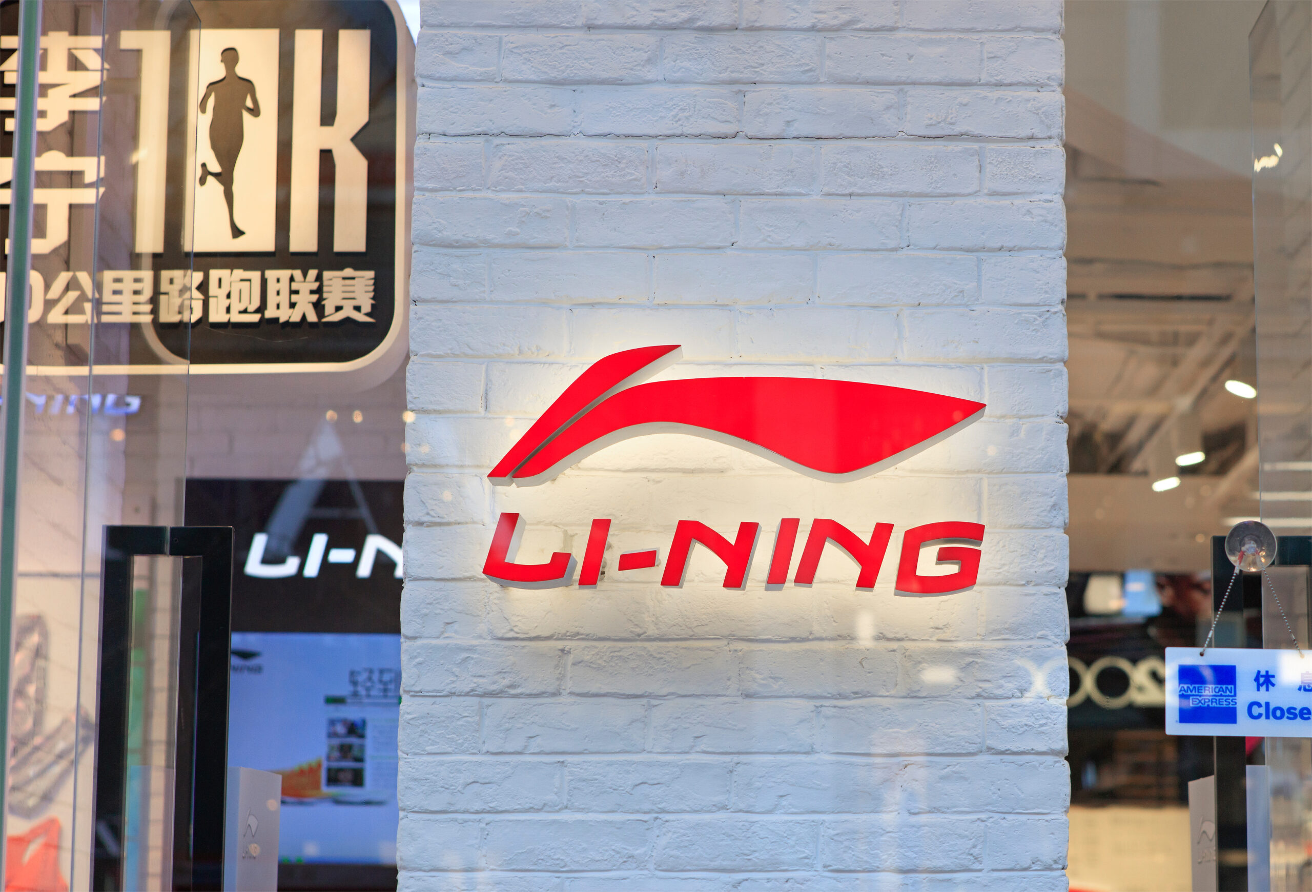 Storefront sign for Li-Ning on a white brick wall with a red swoosh and glass windows behind it.