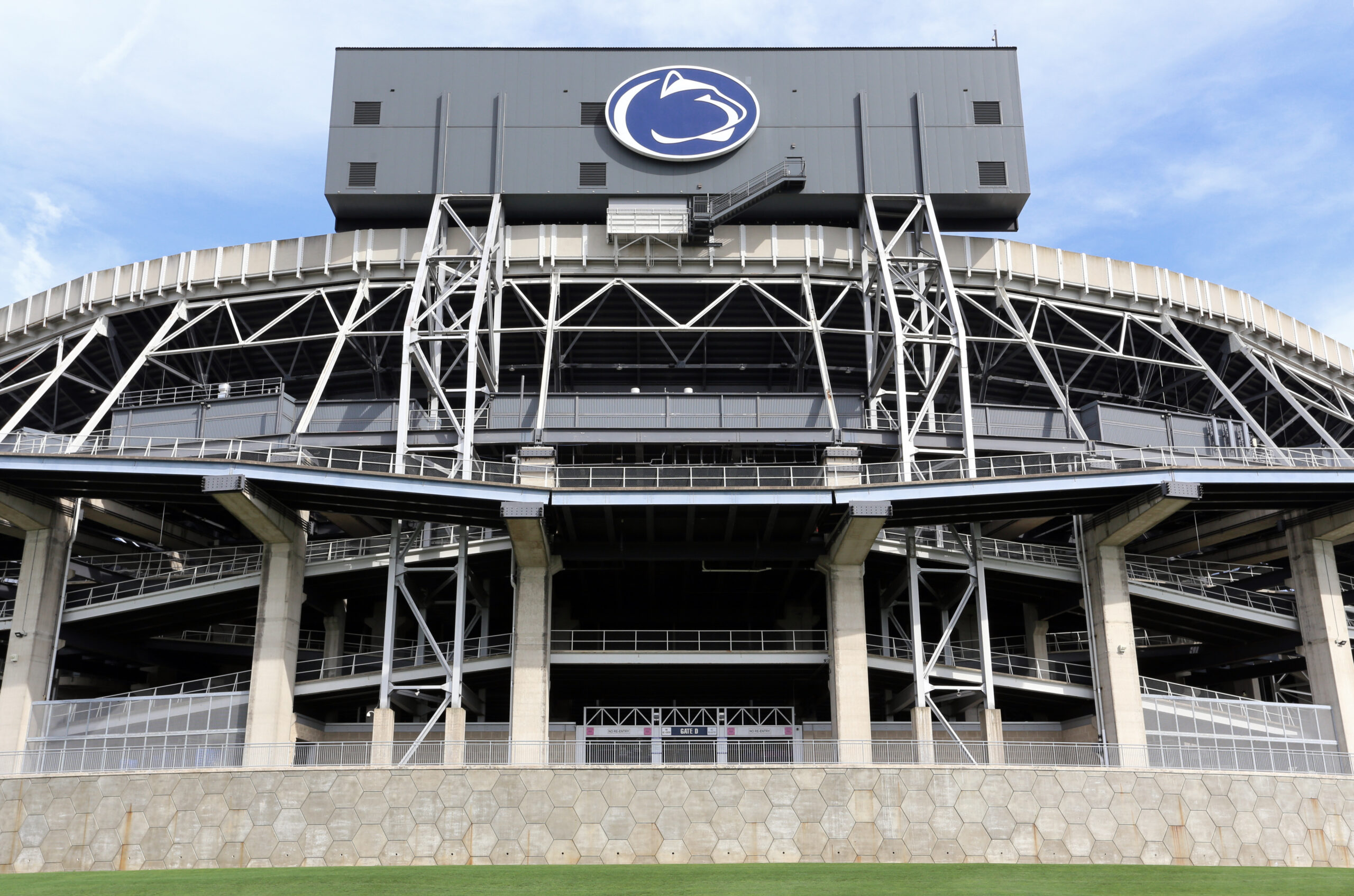 Wide view of a modern stadium exterior with steel framework and a circular blue logo on a gray building against a blue sky, taken from near ground level at the entrance