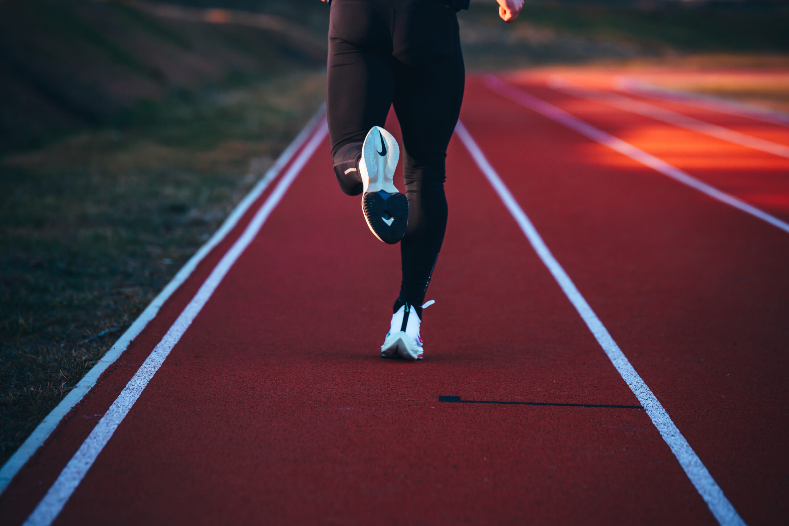 Person running on a red outdoor track, shown from the mid-calf down, wearing black leggings and white sneakers with a blue Nike logo.