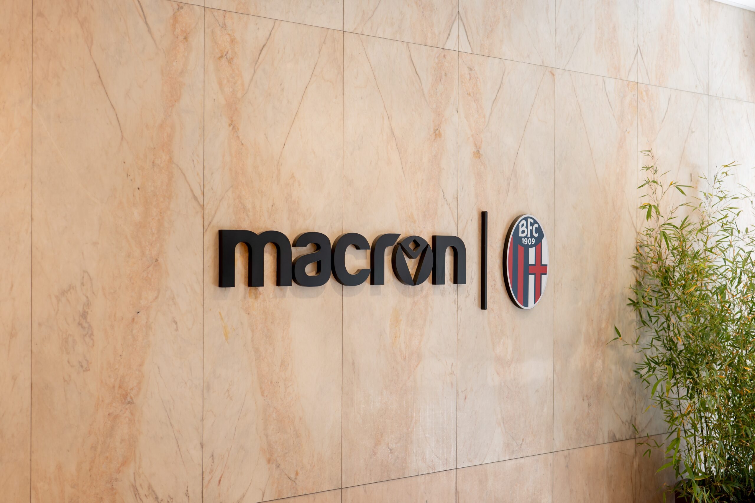 Beige marble wall with a black Macron sign and a round crest logo to the right, near green plants.