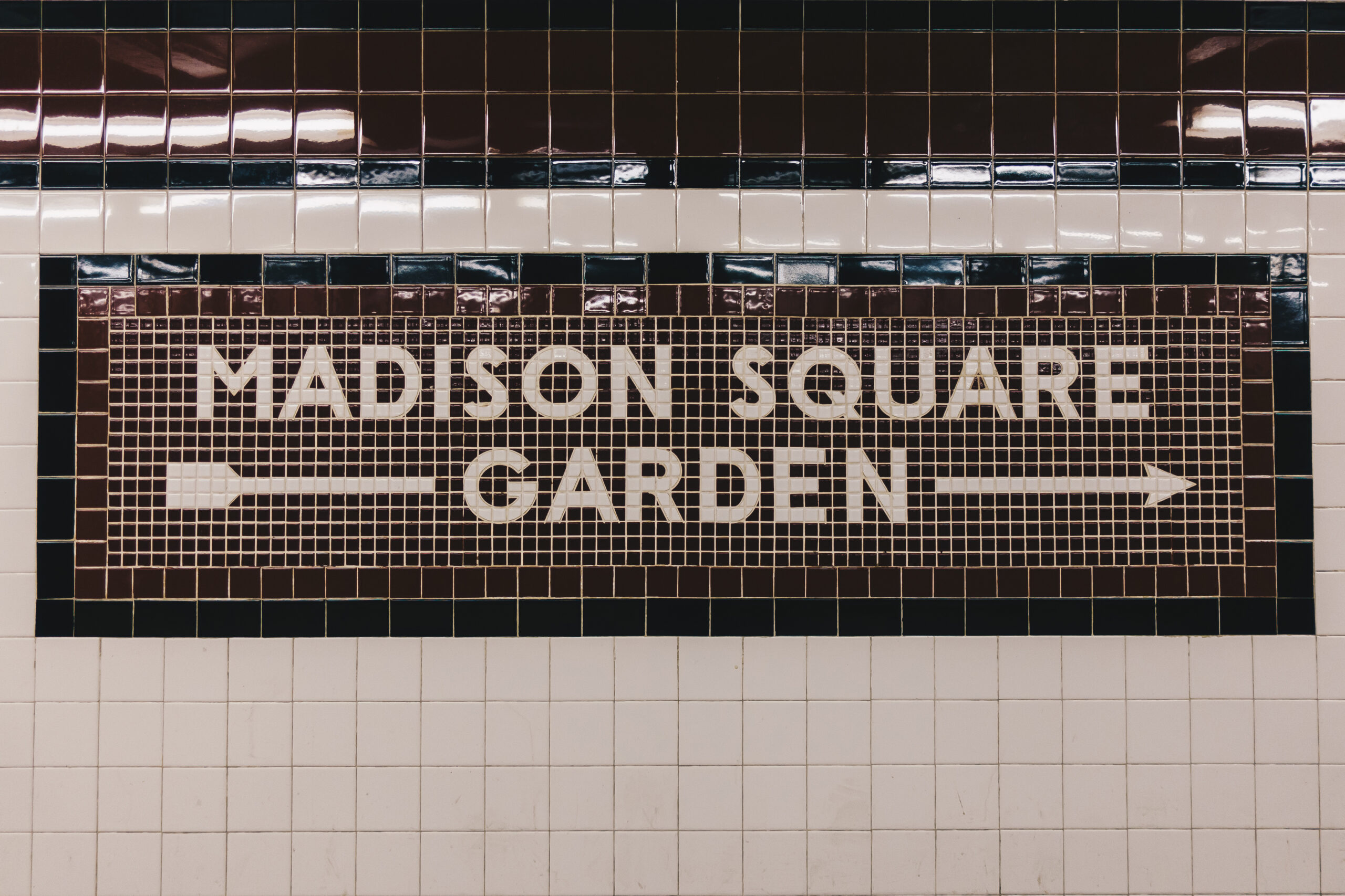 Mosaic tile sign reading 'MADISON SQUARE GARDEN' with an arrow on a tiled wall (brown, black, and beige tiles).