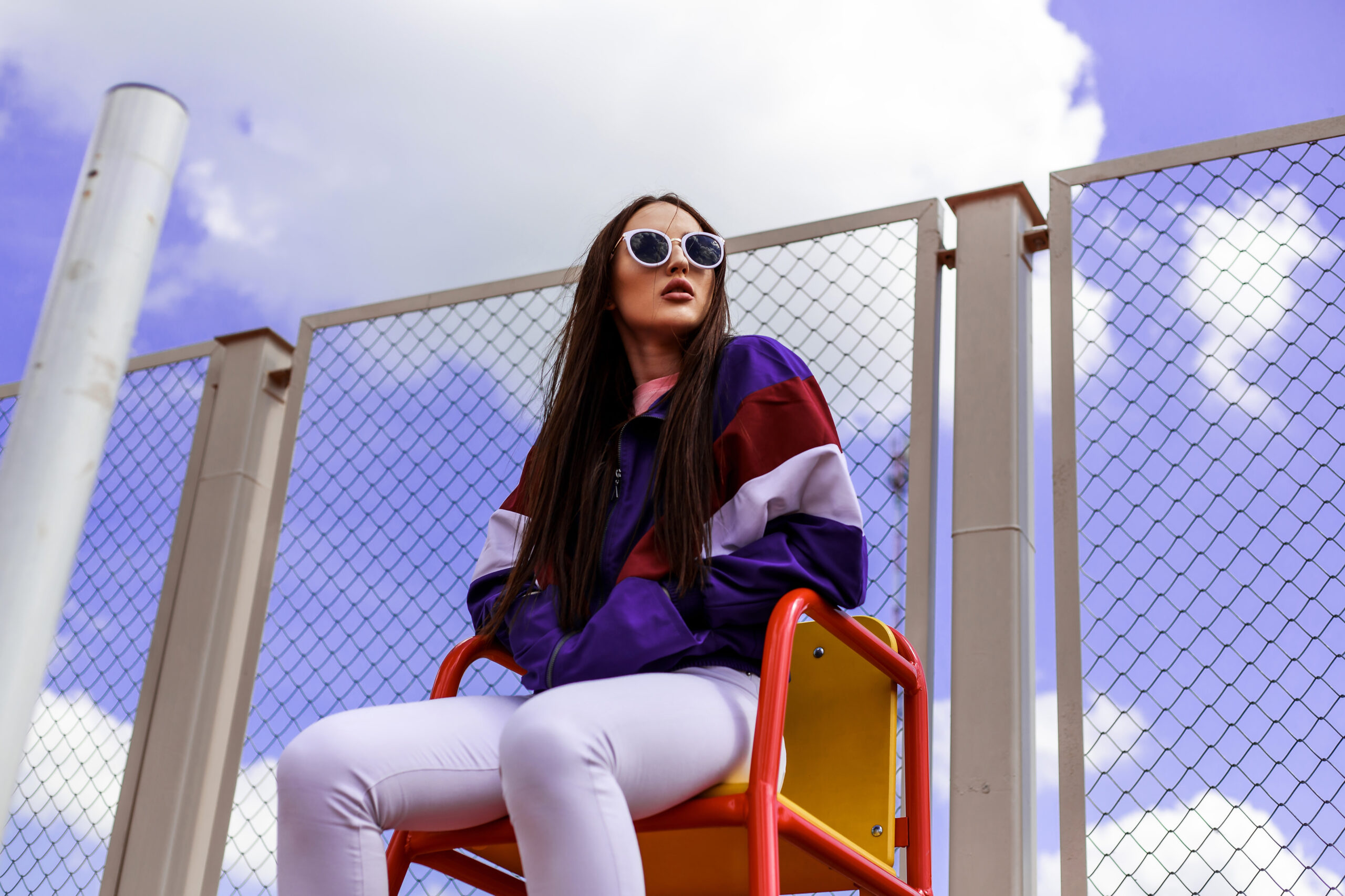 Woman in a purple jacket and white pants sits on a red playground seat beside a chain-link fence with a blue sky behind.