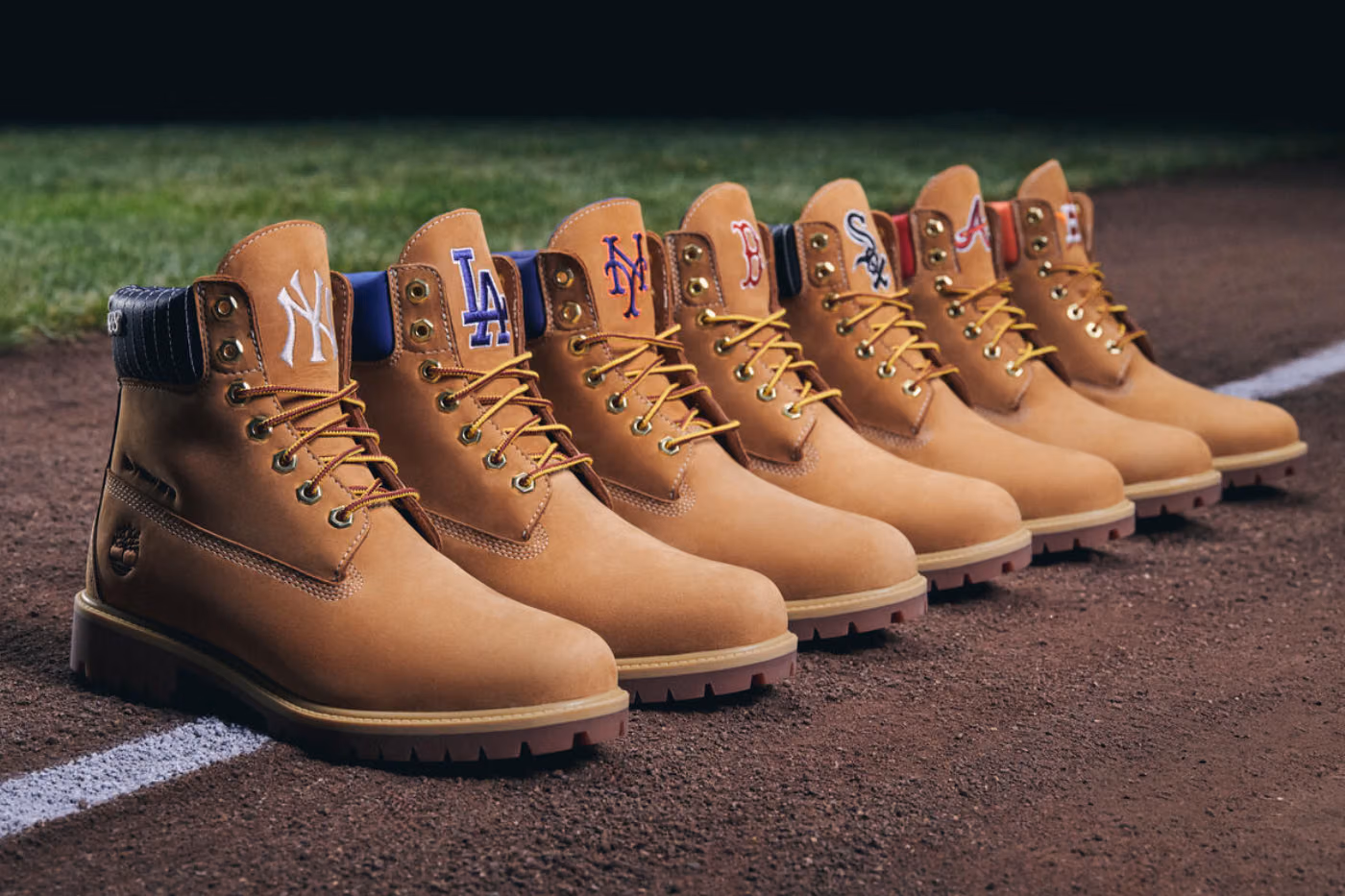 Row of tan boots lined up on a baseball infield, each boot's tongue displaying a different MLB team logo (Yankees, Dodgers, Mets, etc.).